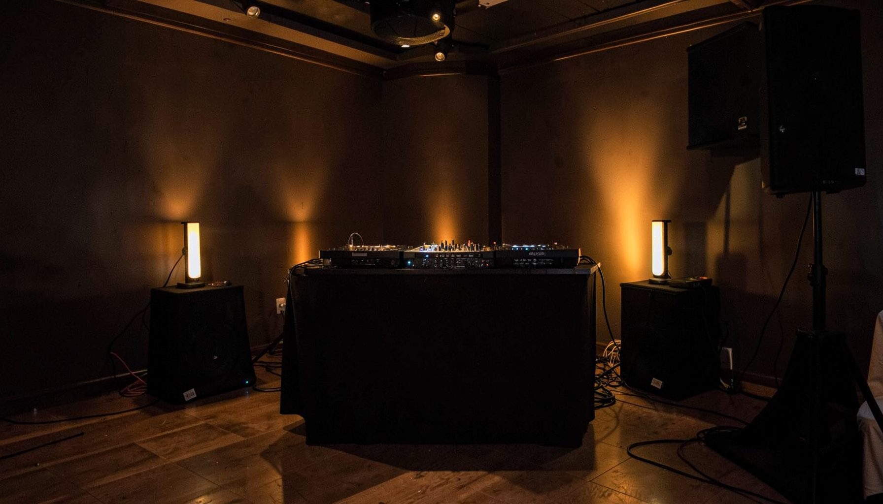 A warm-lit DJ booth setup with speakers against dark wood-paneled walls in an intimate room