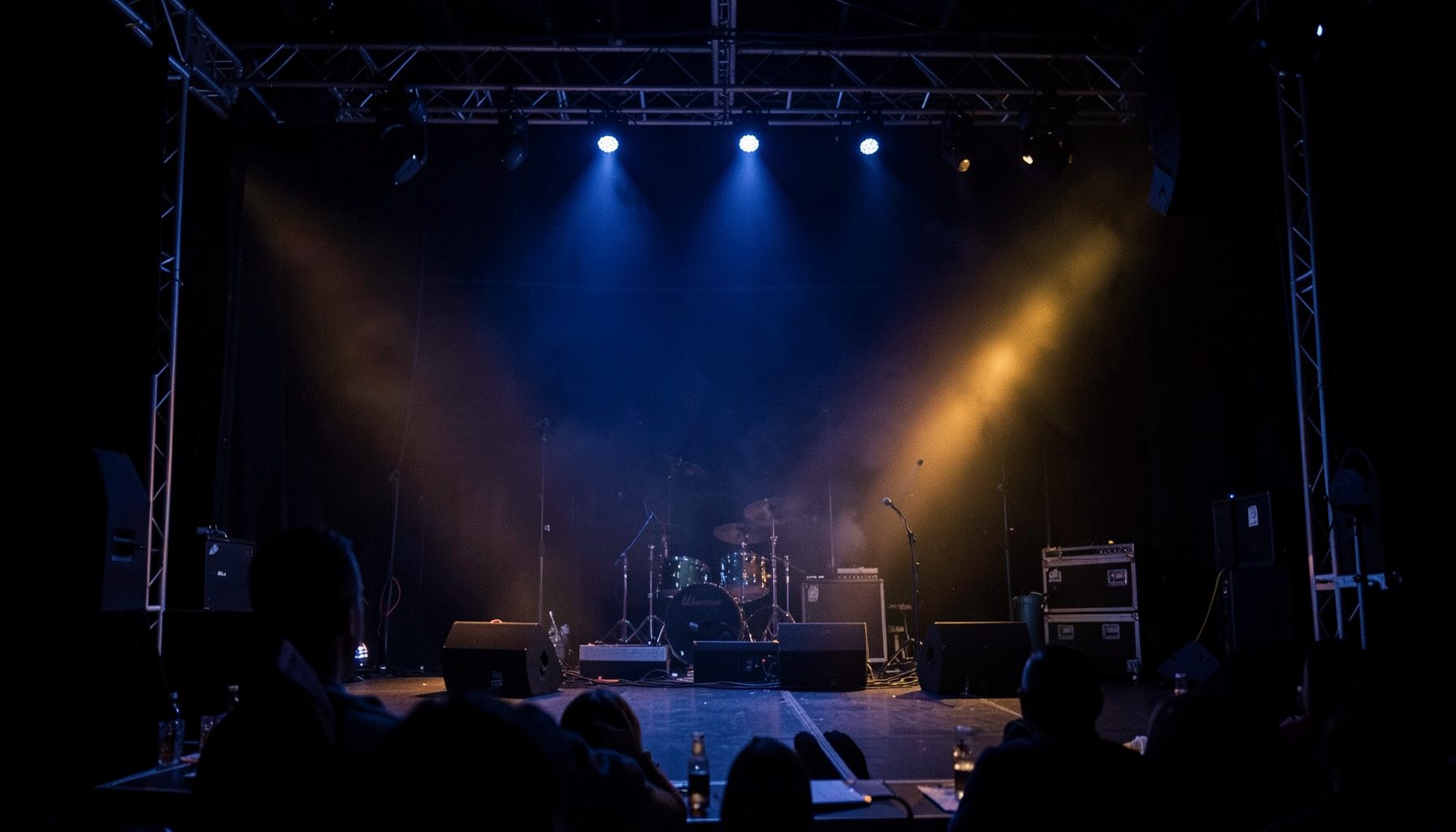 An empty concert stage with drum kit and speakers, lit by blue and amber spotlights in a dark venue